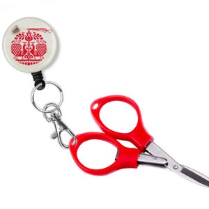 May include: A red pair of small scissors with a retractable cord and a white round keychain with a red embroidered design of two peacocks.