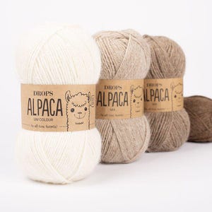 Może przedstawiać: Zbliżenie na kilka motków przędzy Drops Alpaca w różnych neutralnych kolorach. Etykiety zawierają napisy "UNI COLOUR" i "MIX". Przędza to miękkie włókno naturalne, idealne do robótek na drutach i szydełku.