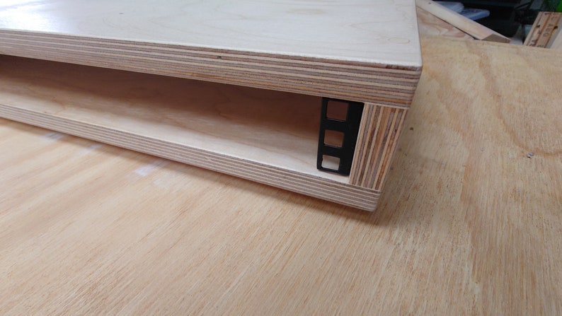 May include: A wooden shelf unit with a single shelf and a black metal rack mounted on the side. The shelf is made of plywood and has a natural wood finish.