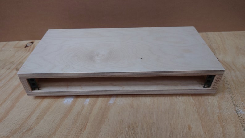 May include: A light wood shelf unit with a single shelf. The shelf is made of plywood and has a natural finish. The shelf is supported by black metal brackets.