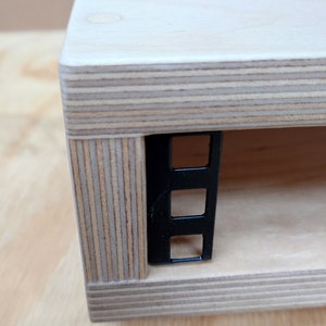 May include: Close-up of a wooden shelf with a black metal rack mounted on the side. The shelf is made of plywood with visible layers of wood veneer.