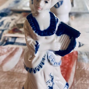 Könnte beinhalten: Eine Porzellanfigur einer Person mit breitkrempigem Hut und einem langen Kleid mit Rüschenbesatz, die eine Harfe hält. Die Figur ist mit einer glänzenden cremefarbenen Glasur überzogen und weist dunkelblaue Akzente am Kragen, Hut und Saum auf.