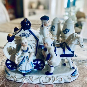May include: A white and blue porcelain figurine set featuring a horse-drawn carriage with a driver and a passenger standing beside it. The carriage has ornate blue and gold detailing, and the figures are dressed in period-style clothing.