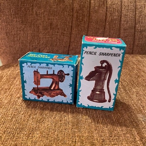May include: Two die-cast miniature pencil sharpeners in boxes. One box features a sewing machine, the other a water pump. Both boxes are teal with white trim and the words "Pencil Sharpener".