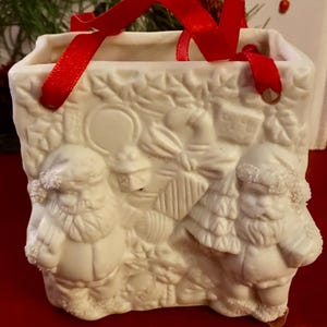 May include: A cream-colored ceramic Christmas ornament shaped like a small tote bag. The ornament features embossed images of Santa Claus, a Christmas tree, and other holiday elements. A red ribbon is attached for hanging.
