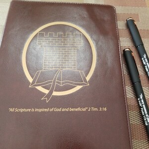 May include: A brown leather-bound journal with a gold embossed design of a castle and open book. The text "All Scripture is inspired of God and beneficial" 2 Tim. 3:16 is printed on the front cover.