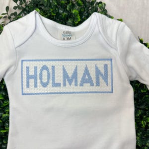 May include: White long-sleeve baby onesie with blue embroidered lettering that says "HOLMAN" in a rectangular frame.
