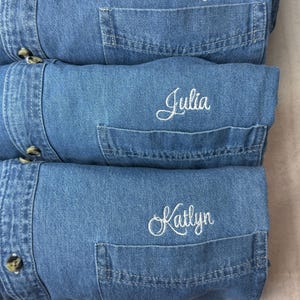 May include: Three folded blue denim shirts stacked vertically. Each shirt features a front pocket with white embroidered script text. The top shirt is embroidered with the name Kailey, the middle with Julia, and the bottom with Katlyn.