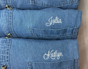 May include: Three folded blue denim shirts stacked vertically. Each shirt features a chest pocket with a name embroidered in white cursive script: Kailey, Julia, and Katlyn. The shirts have visible buttons along the placket on the left side.