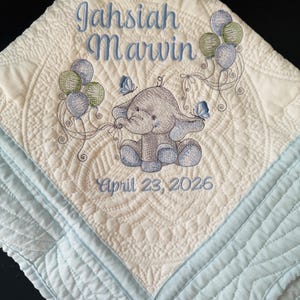 Op de afbeelding: Een gewatteerde babydeken met een geborduurde olifant die ballonnen vasthoudt. De deken is crèmekleurig met blauwe accenten en heeft de naam "Jahsiah Marwin" en de datum "April 23, 2026".
