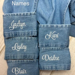 May include: Close-up of a light blue denim shirt with personalized embroidered names in white cursive script on the cuffs. The names include Jaclyn, Lesley, Blair, Kari, and Deidre. The text "Cursive Names" is also visible.