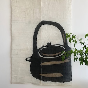 May include: A cream-colored wall hanging with a black and brown illustration of a teapot. The teapot has a large handle and a rounded body. The background has a textured, horizontal pattern.