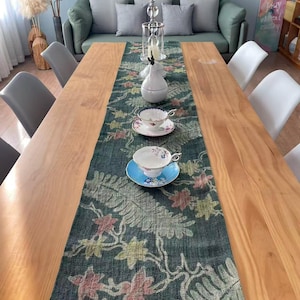 May include: A wooden dining table with a green floral patterned table runner. Two teacups and saucers are placed on the runner. A tall glass candle holder and a white pear-shaped vase are centered on the table. Gray chairs surround the table.