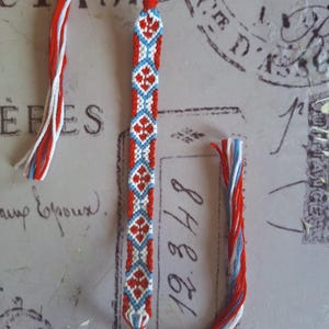 May include: A woven friendship bracelet with a repeating pattern of red, white, and blue diamond shapes. The bracelet is made of threads and has braided ends. The background has faded text and stamps.
