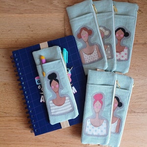 Puede incluir: Varios estuches de lápices azul claro con cremalleras doradas, cada uno con una ilustración única de una mujer. Un estuche contiene bolígrafos y un lápiz. También hay un cuaderno espiral azul.