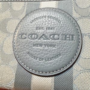 Puede incluir: Una cartera de color azul claro y gris con un patr&oacute;n de logotipo circular repetitivo y rayas verticales. Un parche circular de cuero azul est&aacute; centrado en la parte frontal, grabado con el texto AMERICA'S ORIGINAL EST. 1941 COACH NEW YORK HOUSE OF LEATHER.
