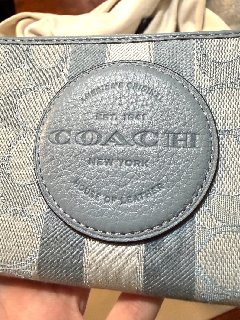 Puede incluir: Una cartera de color azul claro y gris con un patr&oacute;n de logotipo circular repetitivo y rayas verticales. Un parche circular de cuero azul est&aacute; centrado en la parte frontal, grabado con el texto AMERICA'S ORIGINAL EST. 1941 COACH NEW YORK HOUSE OF LEATHER.
