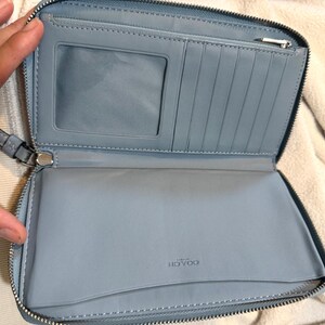 Puede incluir: Una cartera azul claro con cierre de cremallera que presenta un interior con una ventana transparente para identificaci&oacute;n, m&uacute;ltiples ranuras verticales para tarjetas, un bolsillo con cremallera y un compartimento abierto. Marca Coach grabada.