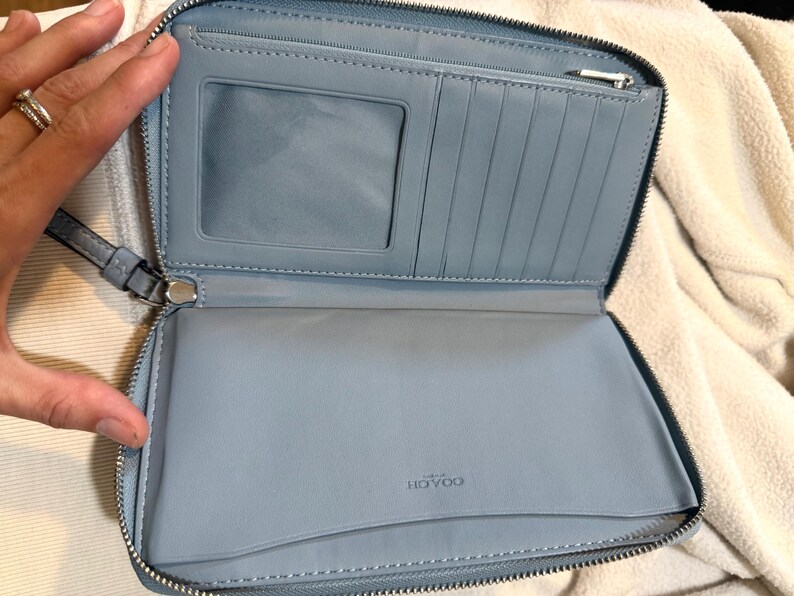 Puede incluir: Una cartera azul claro con cierre de cremallera que presenta un interior con una ventana transparente para identificaci&oacute;n, m&uacute;ltiples ranuras verticales para tarjetas, un bolsillo con cremallera y un compartimento abierto. Marca Coach grabada.