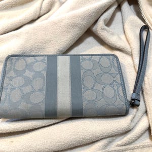 Puede incluir: Una cartera rectangular con correa de mu&ntilde;eca que presenta un patr&oacute;n de logotipo caracter&iacute;stico en azul claro y gris sobre un material tipo lona. El centro tiene un panel de rayas verticales en tonos gris y blanco. Incluye una correa de cuero azul.