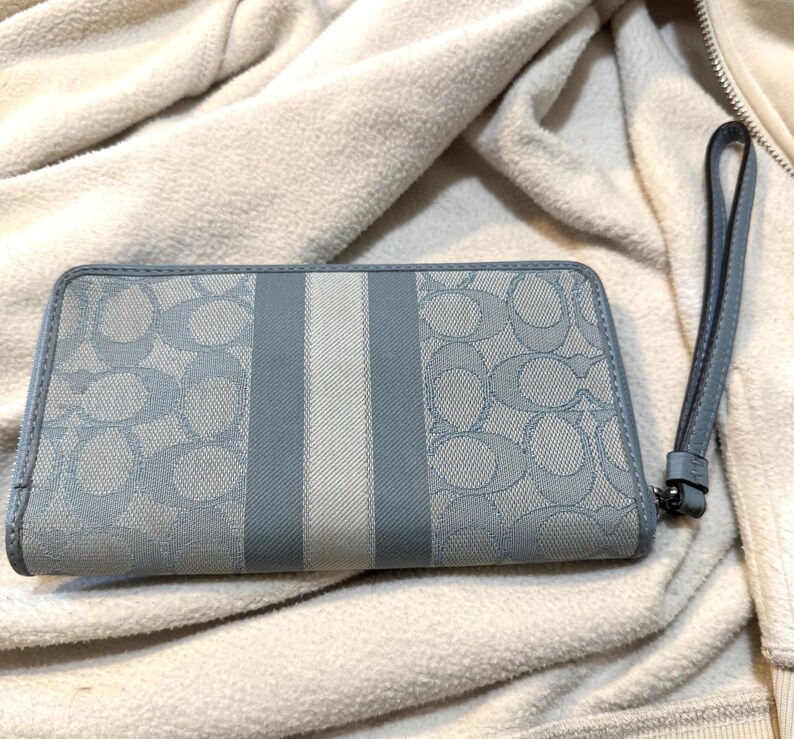 Puede incluir: Una cartera rectangular con correa de mu&ntilde;eca que presenta un patr&oacute;n de logotipo caracter&iacute;stico en azul claro y gris sobre un material tipo lona. El centro tiene un panel de rayas verticales en tonos gris y blanco. Incluye una correa de cuero azul.