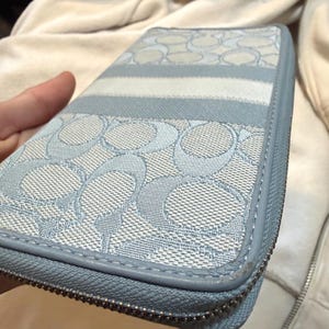 Puede incluir: Una cartera con cremallera en color azul claro y blanco que presenta un patr&oacute;n repetitivo del logotipo C sobre una superficie de tela texturizada. La cartera tiene un borde liso de aspecto cuero azul claro y un cierre de cremallera met&aacute;lico plateado.