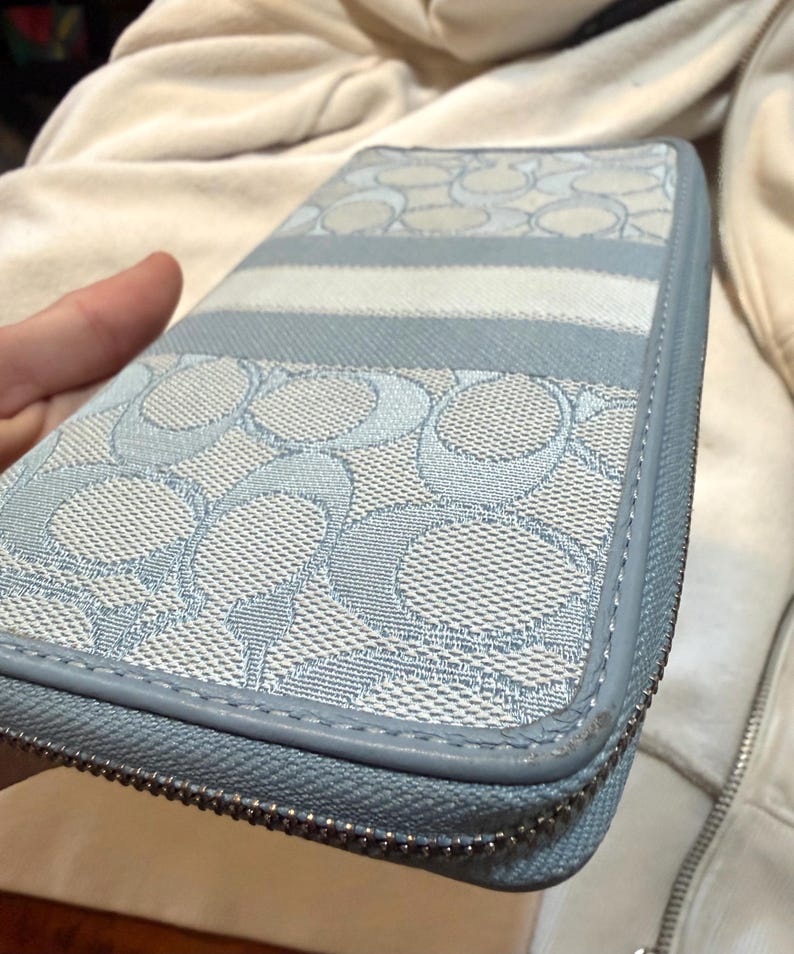 Puede incluir: Una cartera con cremallera en color azul claro y blanco que presenta un patr&oacute;n repetitivo del logotipo C sobre una superficie de tela texturizada. La cartera tiene un borde liso de aspecto cuero azul claro y un cierre de cremallera met&aacute;lico plateado.