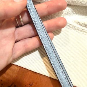 Puede incluir: Una correa de mu&ntilde;eca de color azul claro con una superficie texturizada y granulada, con costuras a juego en los bordes. La correa presenta un acabado de borde liso en azul m&aacute;s oscuro e incluye una anilla de metal de tono plateado.