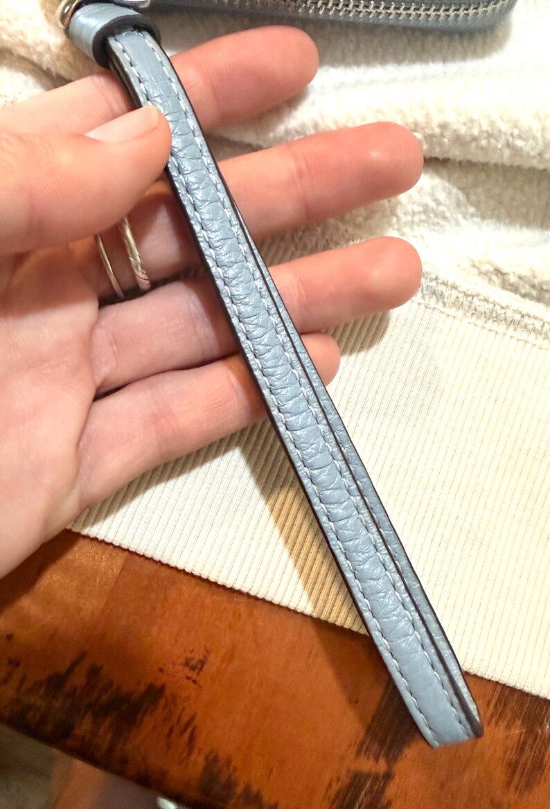 Puede incluir: Una correa de mu&ntilde;eca de color azul claro con una superficie texturizada y granulada, con costuras a juego en los bordes. La correa presenta un acabado de borde liso en azul m&aacute;s oscuro e incluye una anilla de metal de tono plateado.