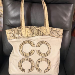 May include: A beige leather tote bag featuring two top handles and a snakeskin-patterned trim along the upper edge. The front of the bag displays a large, centered logo consisting of four interconnected circles with a snakeskin-textured finish.