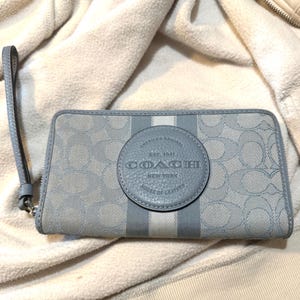 Puede incluir: Una cartera con cremallera en azul claro y gris con un patr&oacute;n de logotipo repetitivo y un parche central de cuero circular con el texto grabado COACH NEW YORK EST. 1941 AMERICA'S ORIGINAL HOUSE OF LEATHER, junto con una correa de mu&ntilde;eca.
