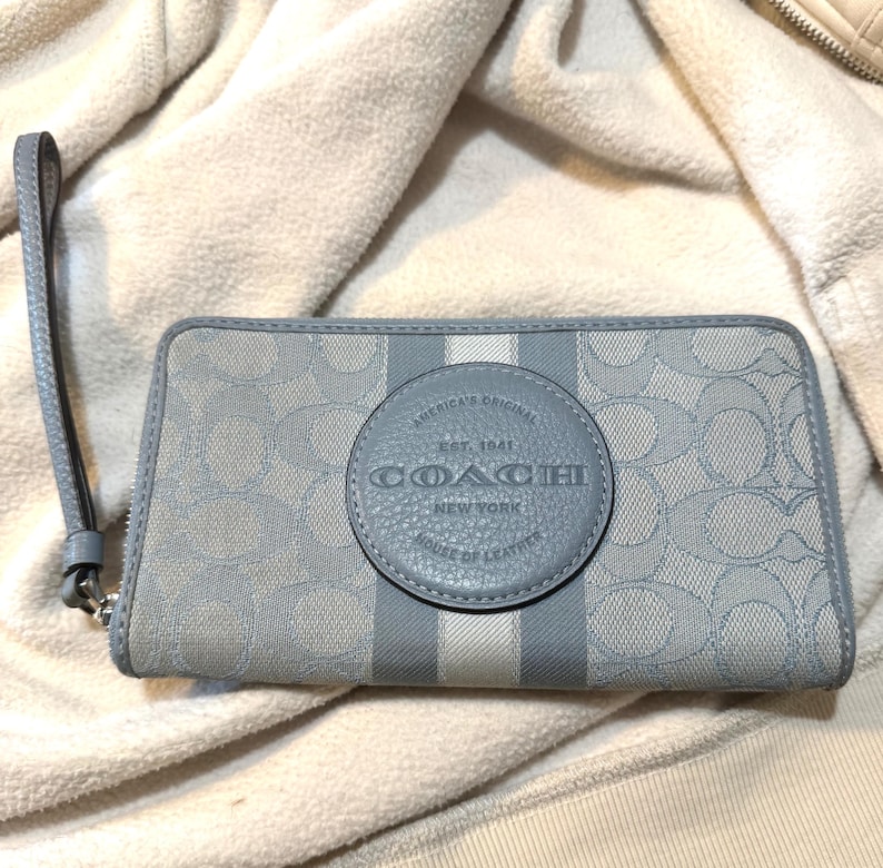 Puede incluir: Una cartera con cremallera en azul claro y gris con un patr&oacute;n de logotipo repetitivo y un parche central de cuero circular con el texto grabado COACH NEW YORK EST. 1941 AMERICA'S ORIGINAL HOUSE OF LEATHER, junto con una correa de mu&ntilde;eca.