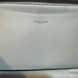 Puede incluir: Una cartera de cuero azul claro con una cremallera oscura en el borde superior. El interior est&aacute; forrado con cuero azul claro a juego y muestra el texto en relieve COACH NEW YORK centrado en el panel superior sobre una ranura horizontal.