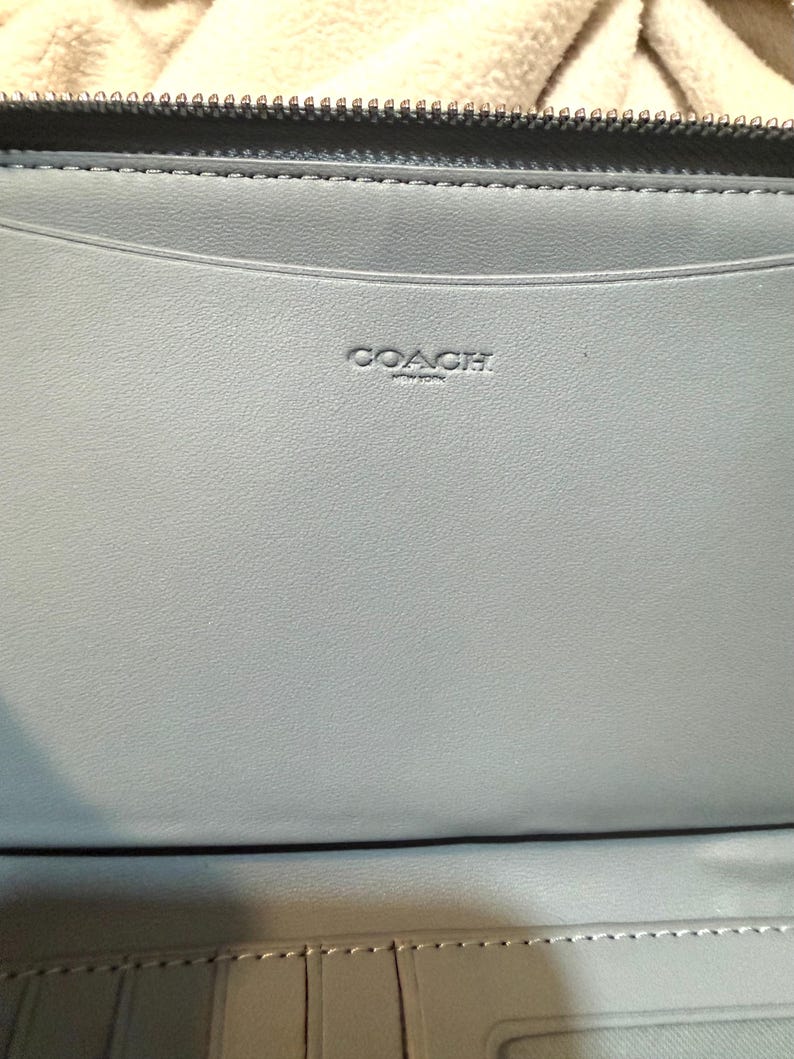 Puede incluir: Una cartera de cuero azul claro con una cremallera oscura en el borde superior. El interior est&aacute; forrado con cuero azul claro a juego y muestra el texto en relieve COACH NEW YORK centrado en el panel superior sobre una ranura horizontal.