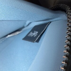 Puede incluir: El interior de una cartera de color azul claro con una superficie texturizada y una etiqueta de tela rectangular oscura con texto en blanco. Una cremallera met&aacute;lica con dientes plateados recorre el lado derecho del compartimento abierto.