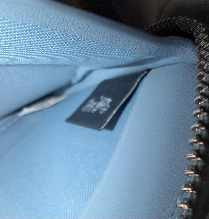 Puede incluir: El interior de una cartera de color azul claro con una superficie texturizada y una etiqueta de tela rectangular oscura con texto en blanco. Una cremallera met&aacute;lica con dientes plateados recorre el lado derecho del compartimento abierto.