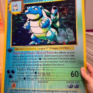 May include: A large, framed Blastoise Pokémon card. The card features a blue and white turtle-like creature with a shell. Text includes "Blastoise", "100 HP", and "Hydro Pump". The card measures 5' 3" in length and weighs 22 lbs.