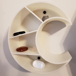 May include: White crescent-shaped shelf with five compartments, each holding a different colored stone. The shelf is mounted on a white wall.