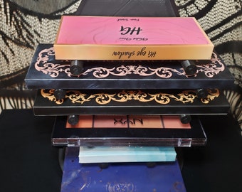 May include: A stack of five eyeshadow palettes in various colors and designs. The top palette is pink with gold lettering that reads "Huda Beauty" and "The New Nude." The second palette is black with gold ornate designs. The third palette is black with a clear plastic cover and the word "NYX" in white letters. The fourth palette is light blue with a white design. The bottom palette is dark blue with gold lettering that reads "The Zodiac."