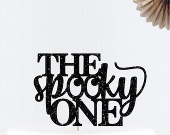 May include: Black glitter cake topper with the text "The spooky one" on a white cake with pink and white flowers.