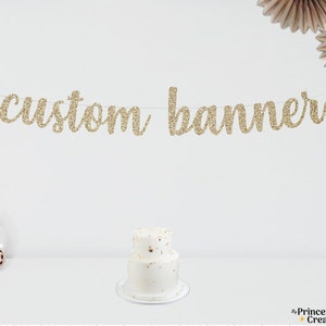 May include: Gold glitter custom banner with the text "custom banner" hanging above a white two-tiered cake.