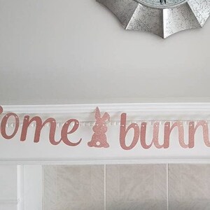 May include: A rose gold glitter banner that reads "Some bunny is one". The banner is hanging on a white mantelpiece with two silver candle holders on either side.
