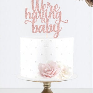 May include: A white cake with a pink glitter cake topper that says "We're having a baby". The cake is on a gold cake stand and has pink and white flowers on top.