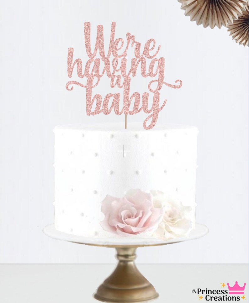 May include: A white cake with a pink glitter cake topper that says "We're having a baby". The cake is on a gold cake stand and has pink and white flowers on top.