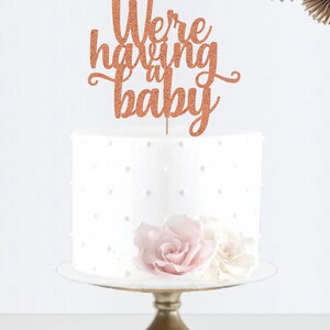 May include: Rose gold glitter cake topper with the text "We're having a baby".
