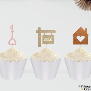 May include: Three cupcakes decorated with gold and pink glitter toppers. The toppers are a key with a heart, a house with a heart, and a sold sign.