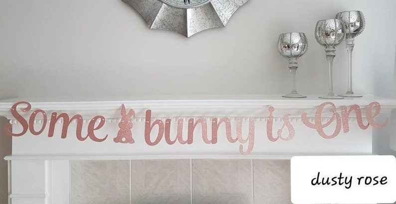 May include: A rose gold glitter banner that reads "Some bunny is one". The banner is hanging on a white mantelpiece with two silver candle holders on either side.