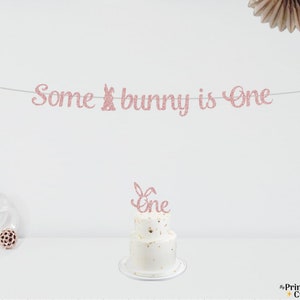 May include: Pink glitter banner with the text "Some bunny is One" and a pink glitter cake topper with the text "One" on a white cake with gold sprinkles.