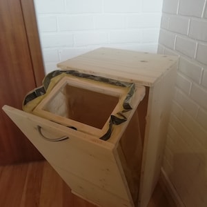 Könnte beinhalten: Ein helles Holz-Mülleimer mit einer Scharniertür und einem Griff. Der Behälter ist rechteckig mit einer schrägen Vorderseite und einem hochklappbaren Deckel. Ein gelber und schwarzer Kunststoffeinsatz ist sichtbar.