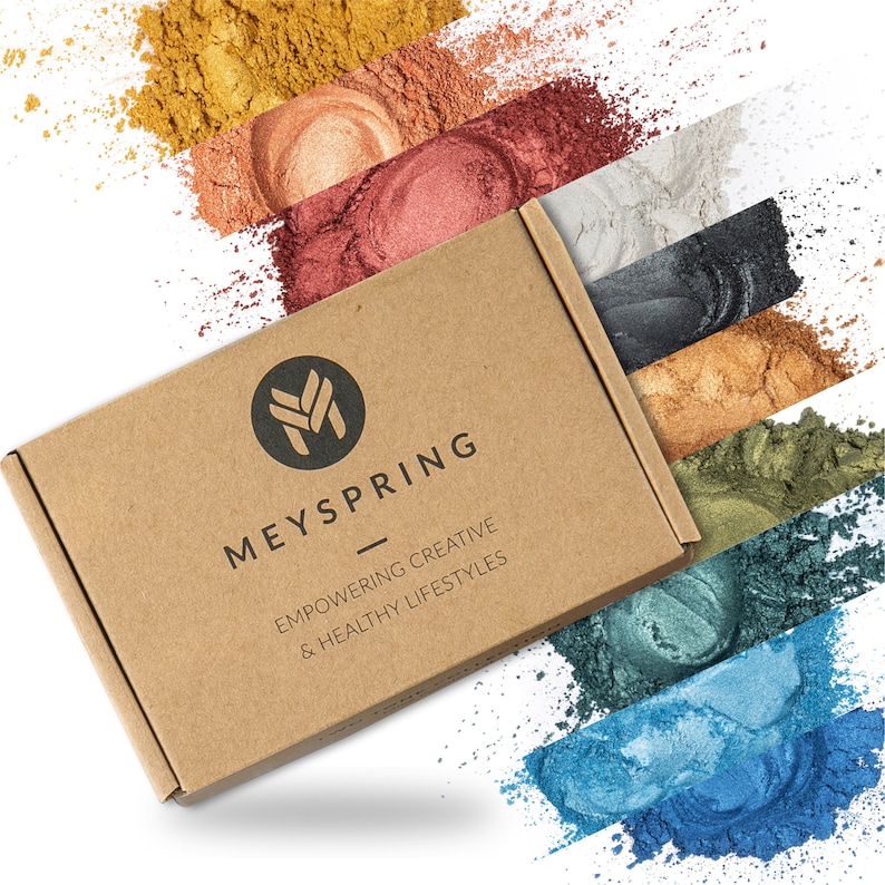 May include: A brown cardboard box with the text "MEYSPRING EMPOWERING CREATIVE & HEALTHY LIFESTYLES" printed on it. The box is surrounded by colorful powder pigments in various shades of gold, red, brown, gray, black, green, and blue.